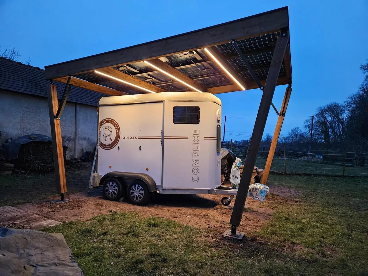 Un carport solaire en bois éclairé par ses LED à la tombée de la nuit, un van garé dessous